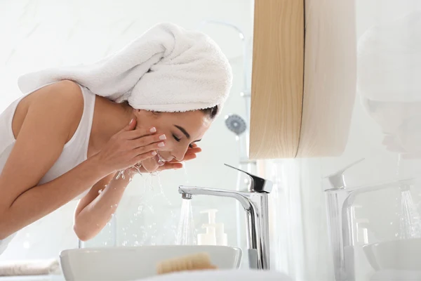 Kobieta w białym ręczniku na głowie myje twarz w łazience.